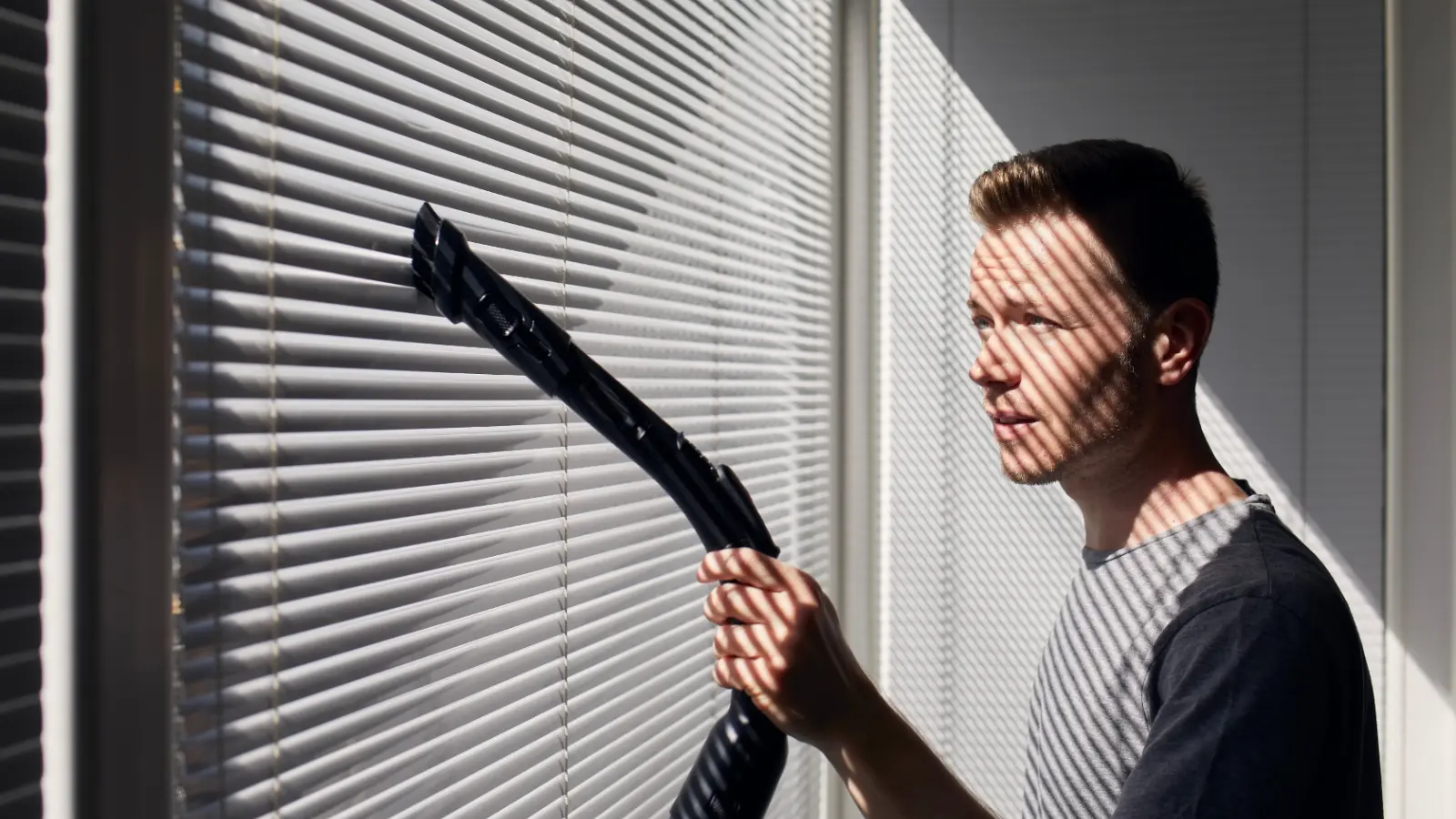 A man cleaning blinds with a vacuum cleaner.