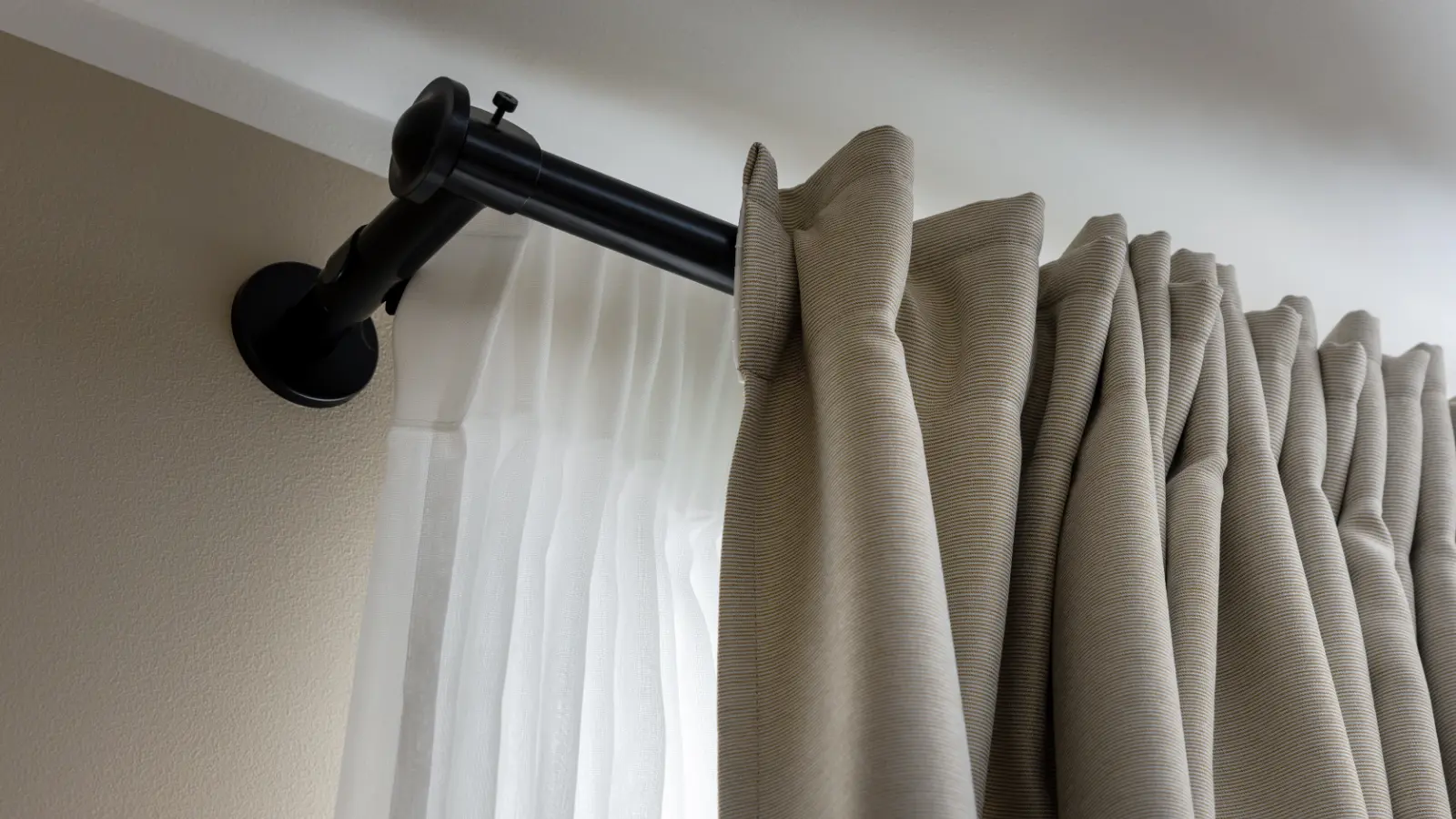 A black curtain rod with beige curtains hanging on it.