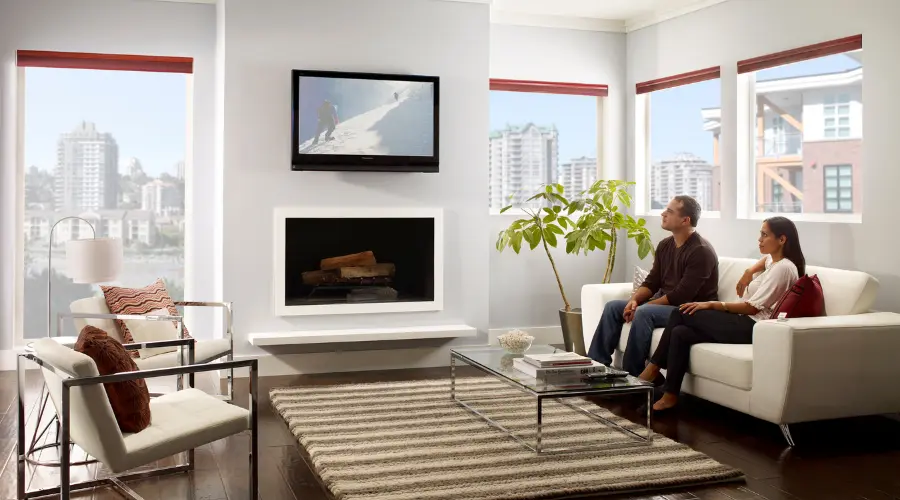 A couple relaxing on a white sofa in a modern living room with large windows, a fireplace, and a mounted television displaying a snowy landscape.