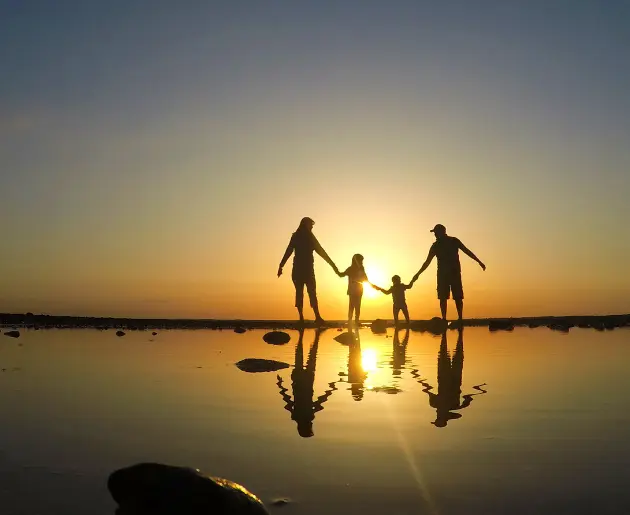 Silhouette of a family of four holding hands against a setting sun on the beach.